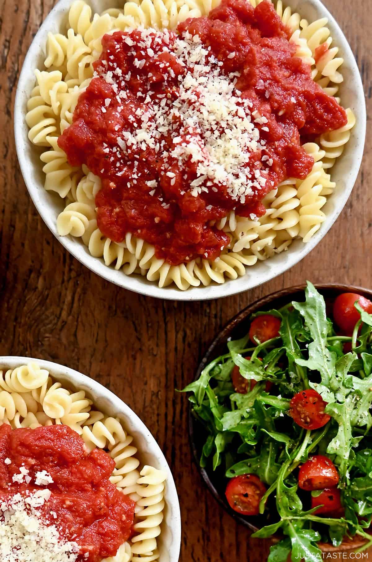 A bowl containing pasta topped with easy homemade marinara sauce garnished with Parmesan cheese near a plate with a fresh green salad.