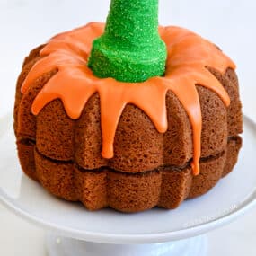 Easy Pumpkin Bundt Cake topped with a thick orange-colored glaze and a green sugar cone "stem" on a cake platter