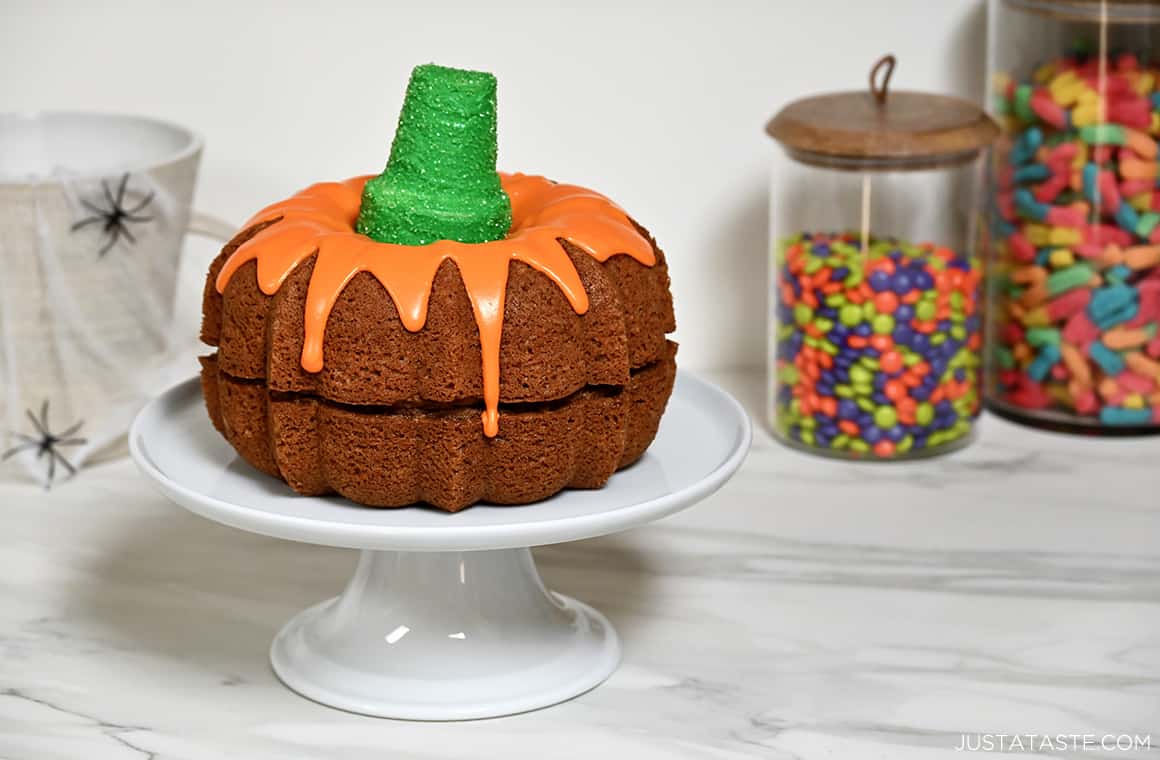 A pumpkin bundt cake with orange vanilla frosting and a green cone "stem" on a cake stand next to glass jars filled with candy