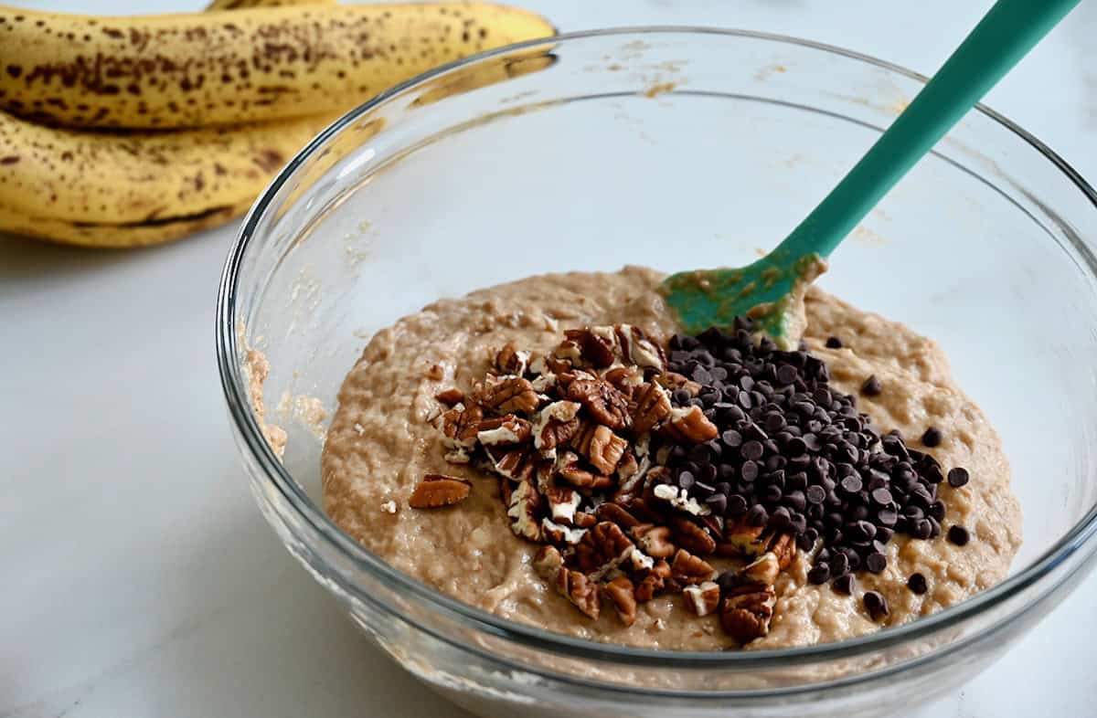 Whole wheat banana bread batter topped with mini chocolate chips and chopped pecans in a glass bowl next to a bunch of overripe bananas.