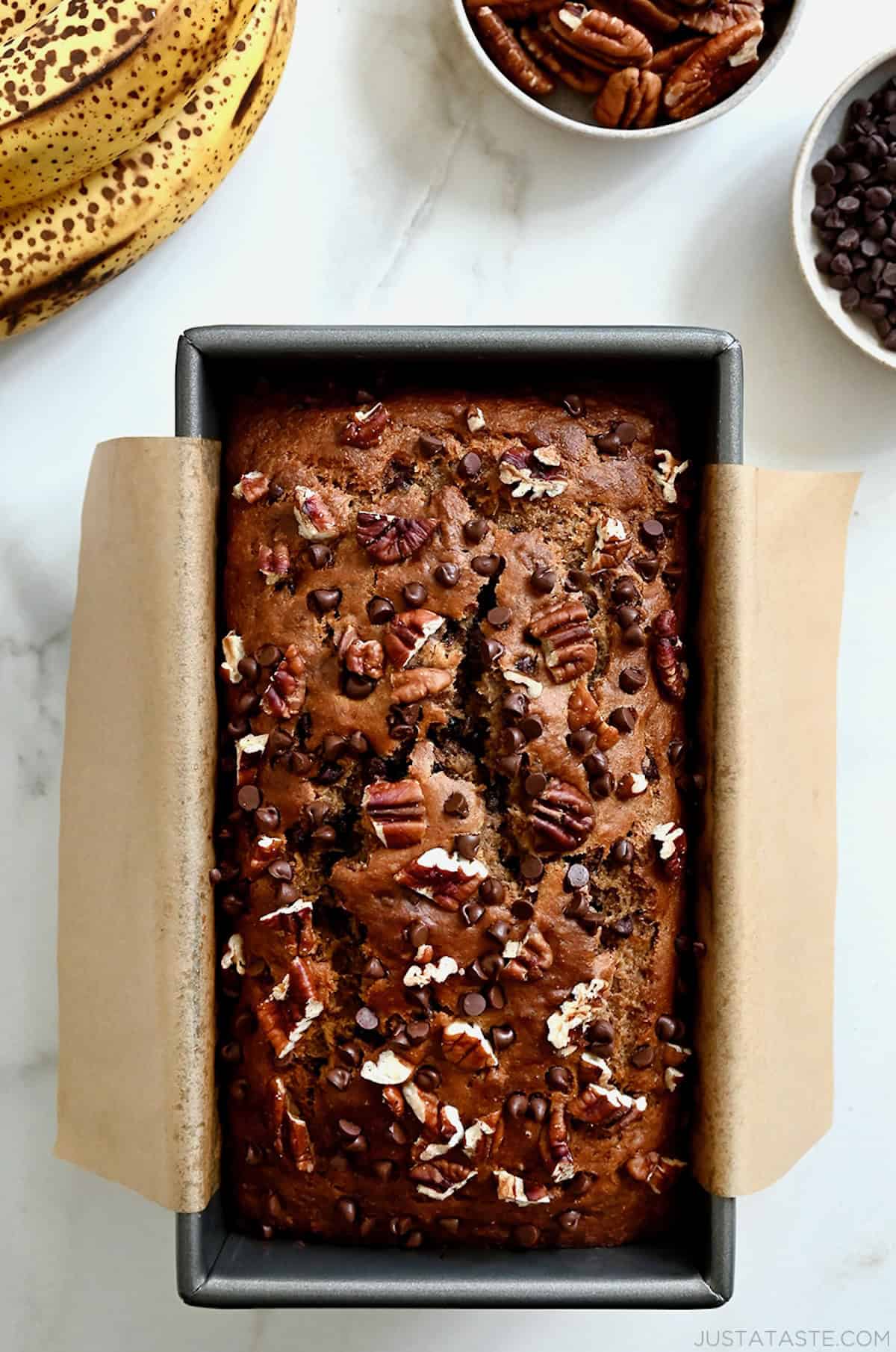 Whole wheat banana bread topped with mini chocolate chips and chopped pecans in a parchment paper-lined bread pan.