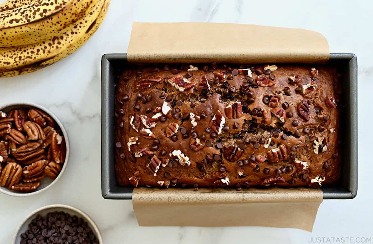 Healthy wheat banana bread in a loaf pan lined with parchment paper next to a bunch of overripe bananas and a small bowl containing mini chocolate chips.
