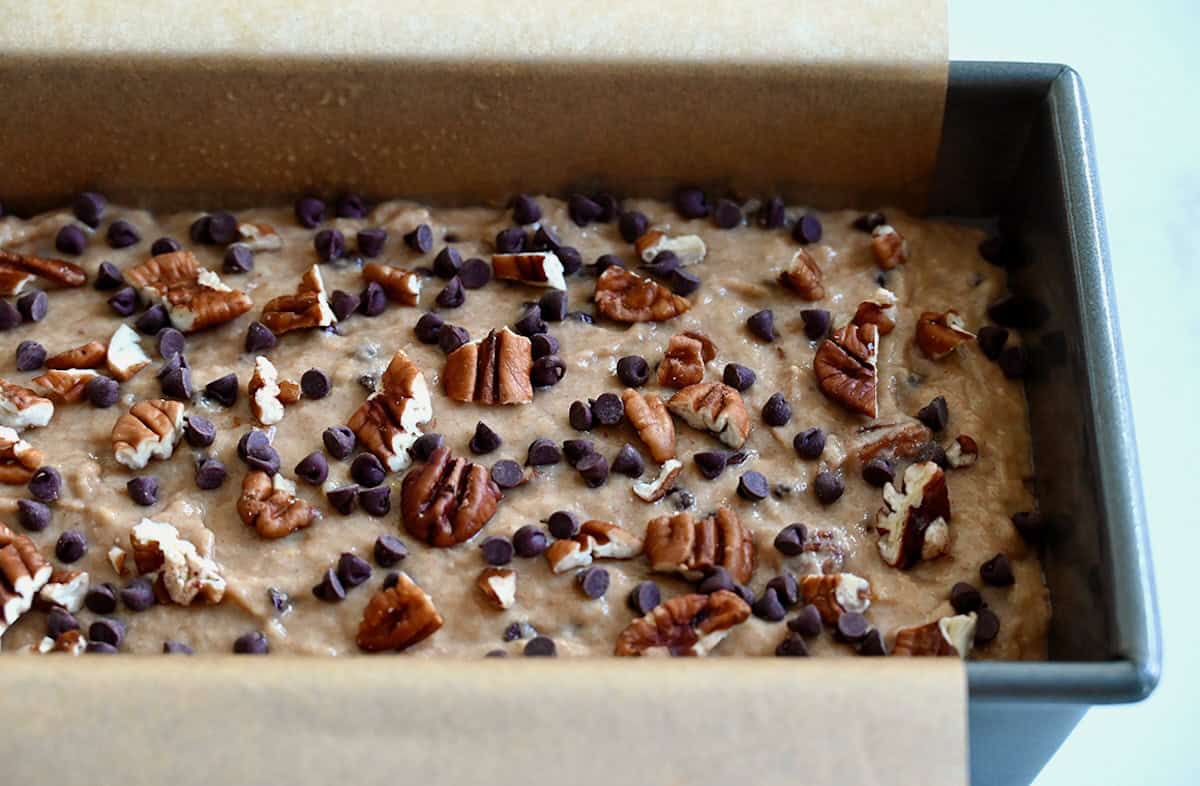 Whole wheat banana bread batter topped with mini chocolate chips and chopped pecans in a parchment paper-lined loaf pan.