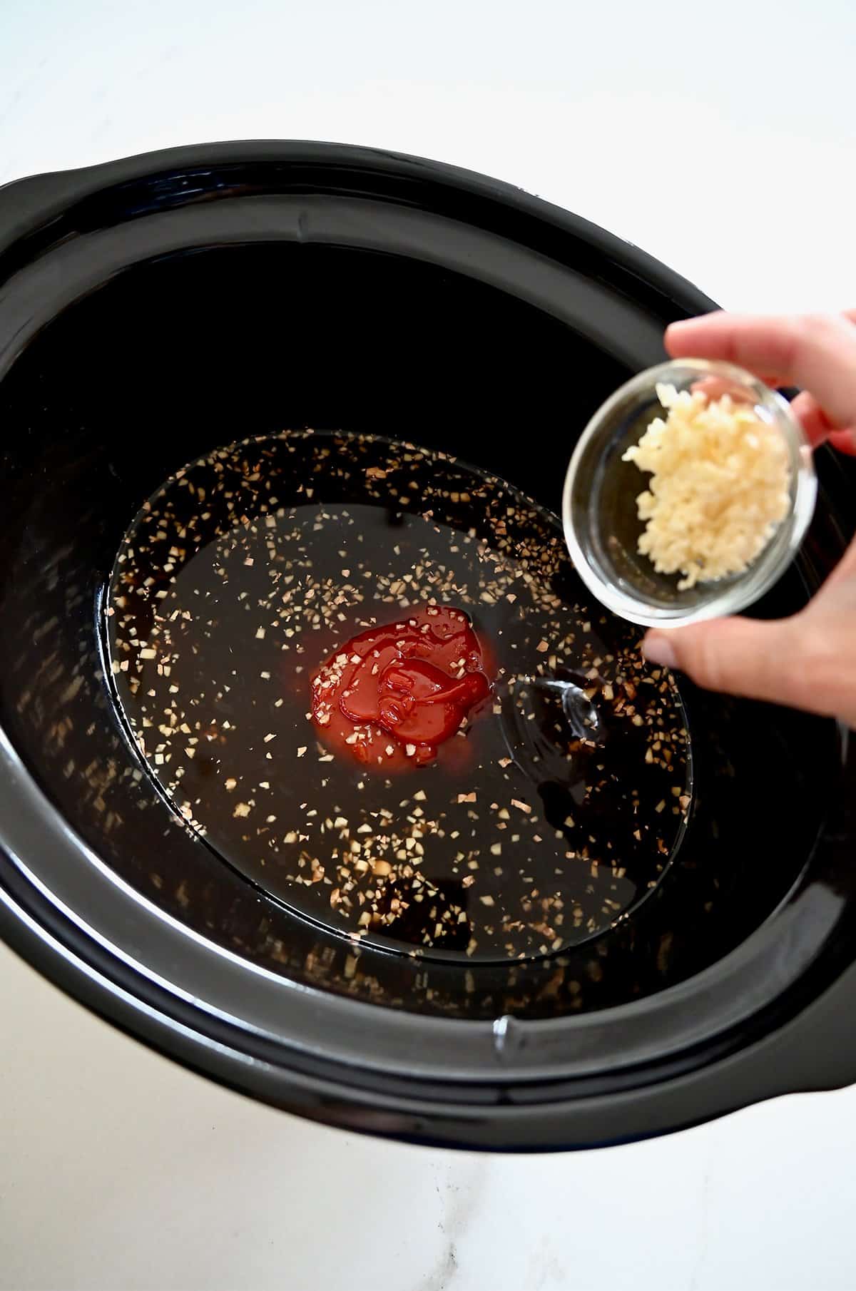 Adding minced garlic and ginger to a slow cooker with soy sauce, chicken stock, pineapple juice, ketchup, and brown sugar.