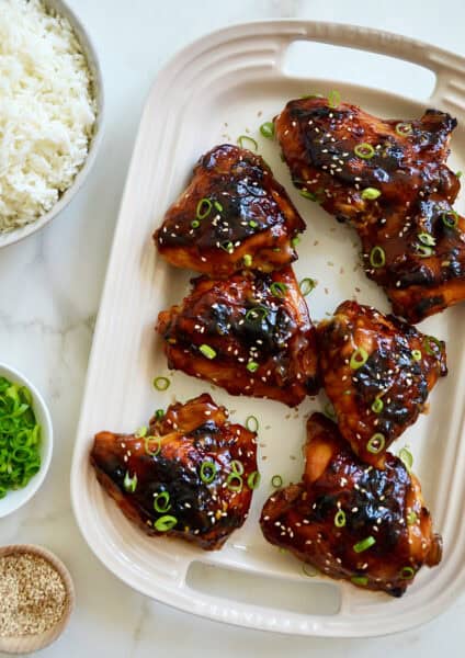Glazed honey soy chicken thighs garnished with sesame seeds and chopped scallions on a white serving platter.