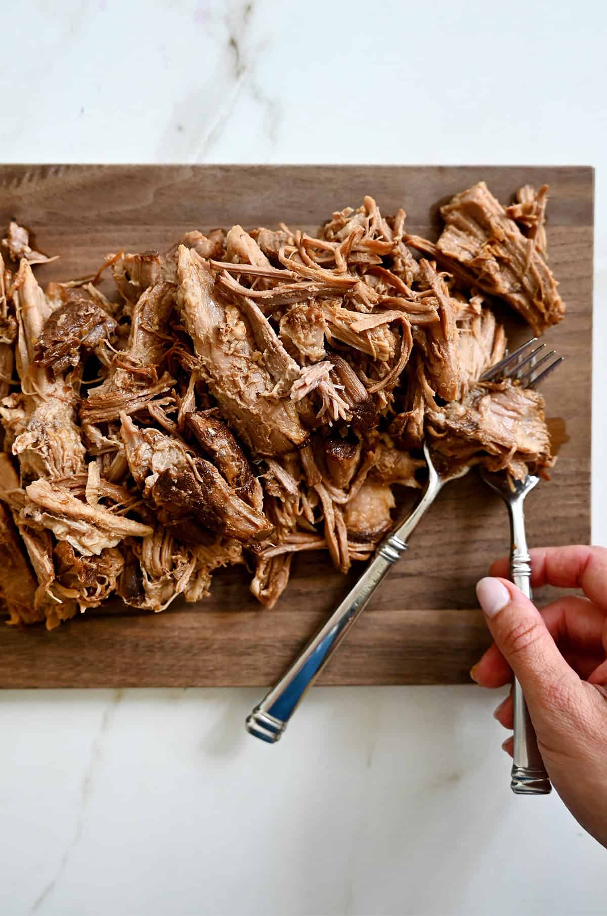 Using two forks to shred slow-cooked pork on a cutting board.