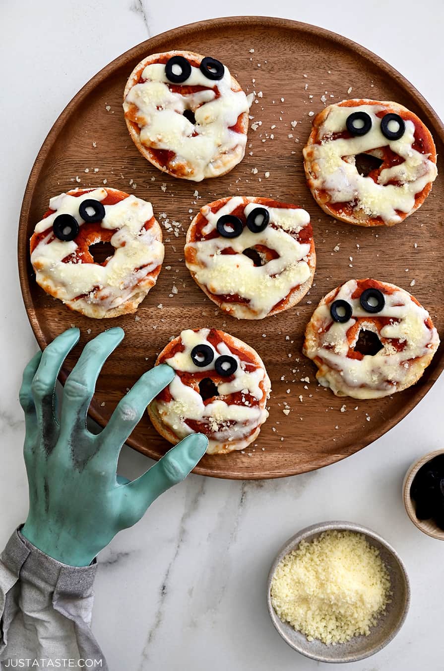 A wood plate containing mini bagel pizzas and a mummy hand