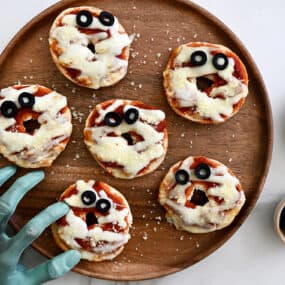A wood platter with mini bagel pizzas and a green mummy hand