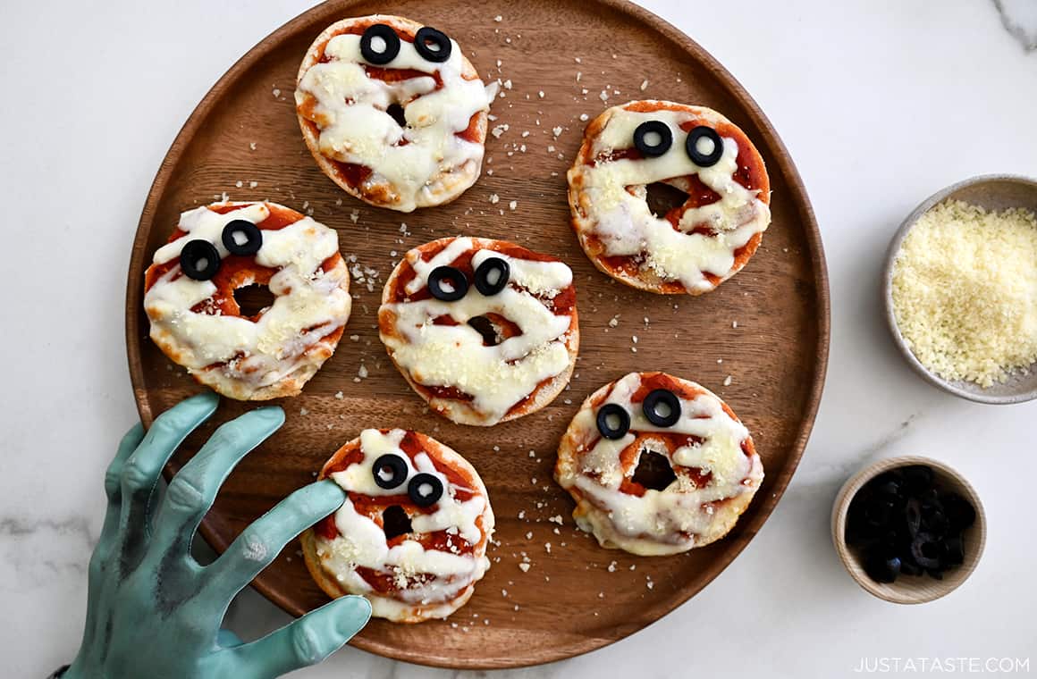 A wood platter with mini bagel pizzas and a green mummy hand