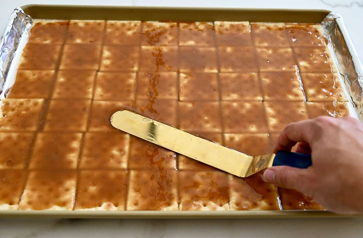 Una mano tiene una spatola offset mentre sparge il caramello sopra i cracker