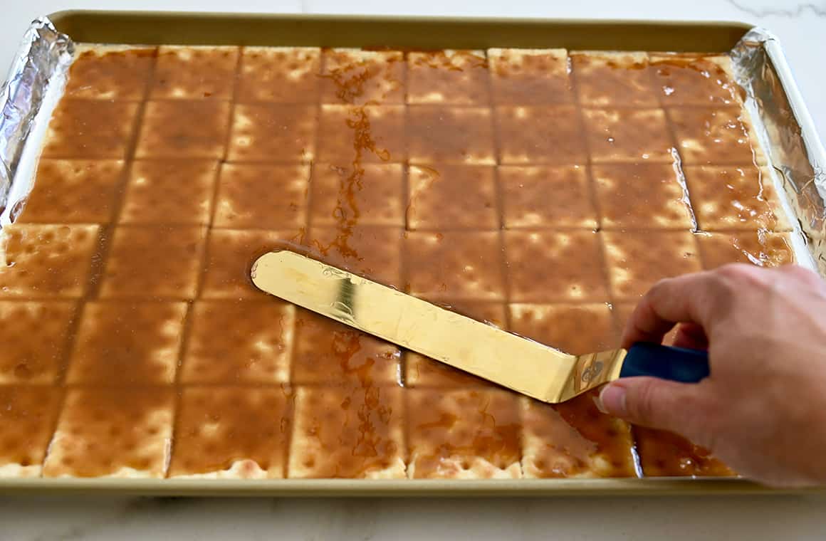 A hand holds an offset spatula while spreading caramel atop crackers