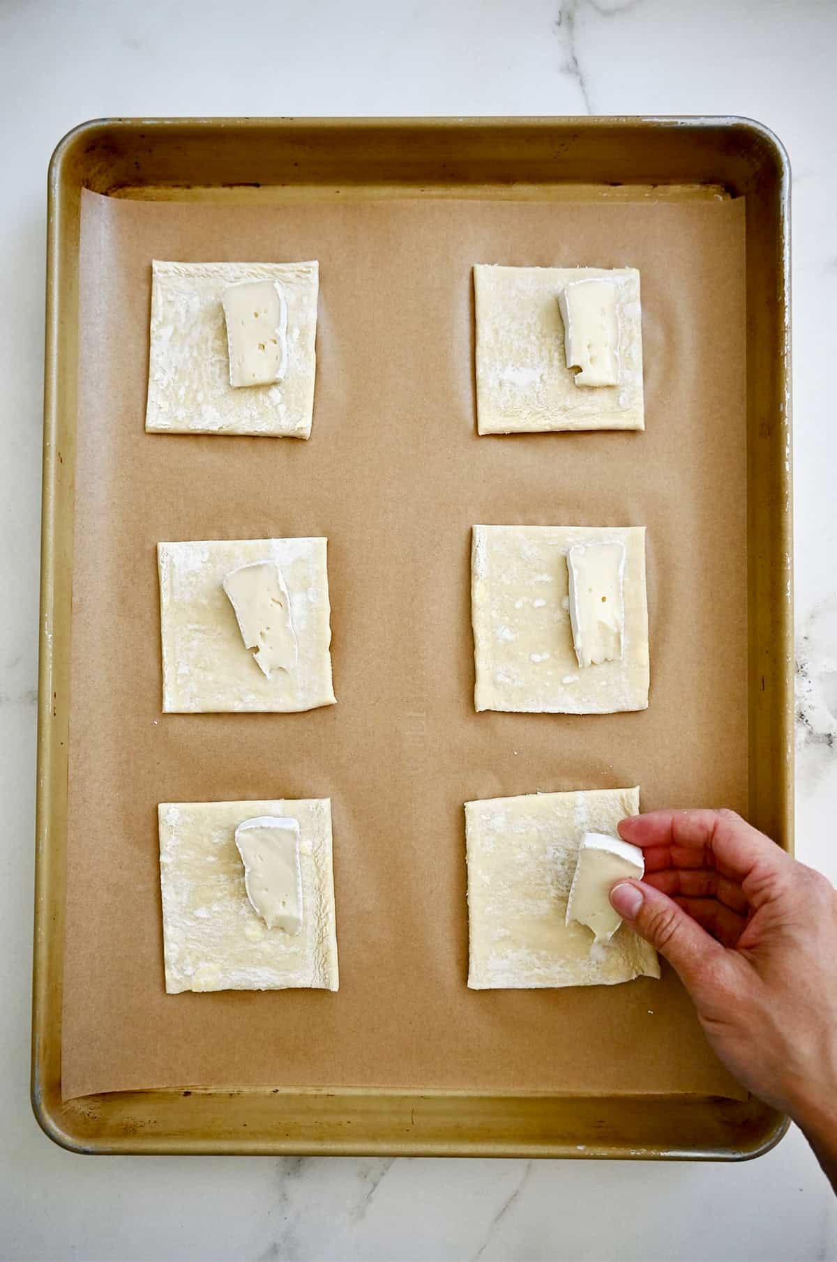 Adding a thin slice of Brie cheese to a square of puff pastry on a parchment paper-lined baking sheet.