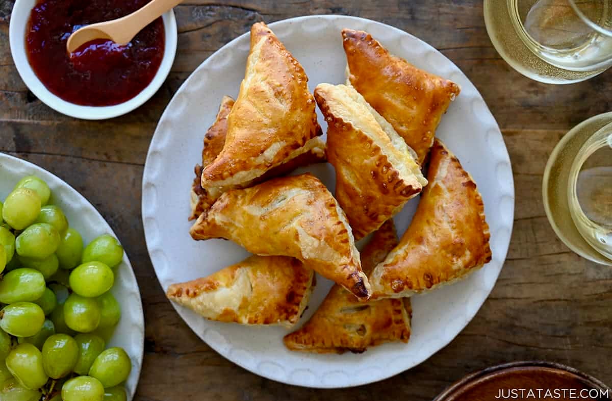 Golden-brown baked brie bites filled with jam on a white plate next to a plate of grapes and two glasses of white wine.