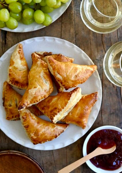 Baked Brie Bites with Jam piled on a plate next to a small bowl containing jam and two glasses of white wine.