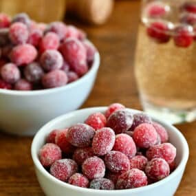 Two white, small bowls containing sugared cranberries next to a glass of champagne with several sugared cranberries