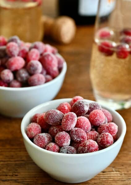 Two white, small bowls containing sugared cranberries next to a glass of champagne with several sugared cranberries