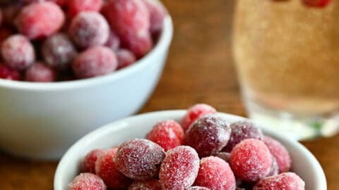 Two white, small bowls containing sugared cranberries next to a glass of champagne with several sugared cranberries