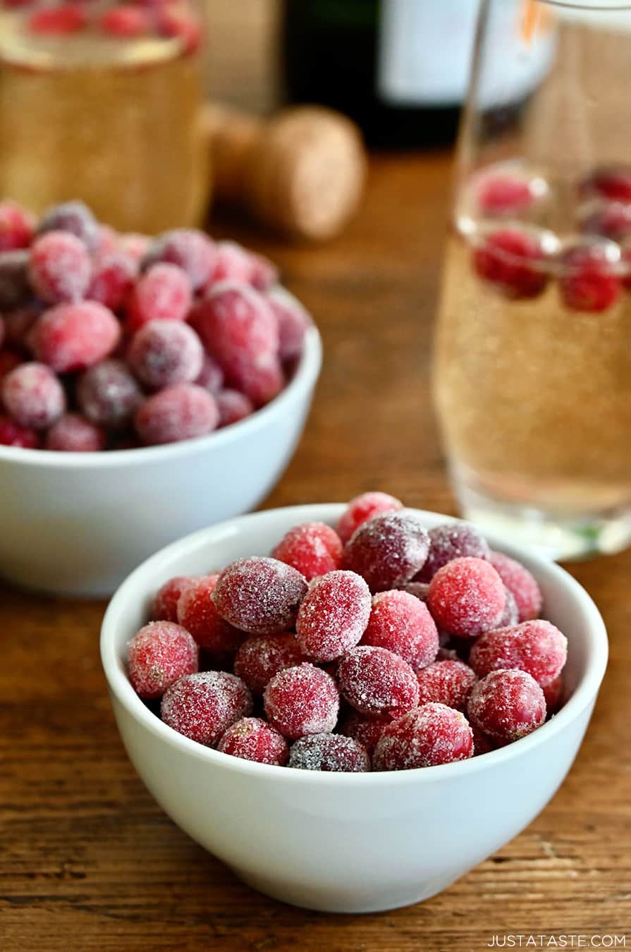 Two white, small bowls containing sugared cranberries next to a glass of champagne with several sugared cranberries