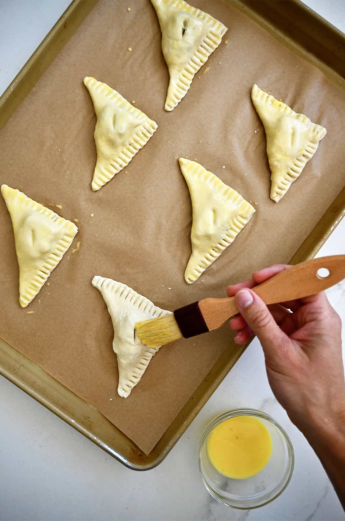 Brushing egg wash onto unbaked puff pastry Brie bites, shaped into triangles and sealed with fork-crimped edges on a parchment-lined baking sheet.