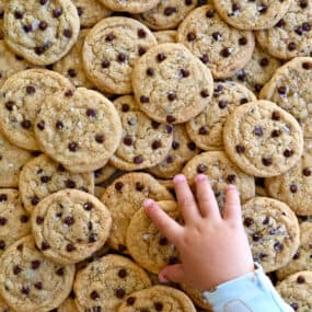 A pile of mini chocolate chip cookies with a child's hand reaching for a cookie.