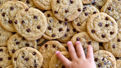 A pile of mini chocolate chip cookies with a child's hand reaching for a cookie.