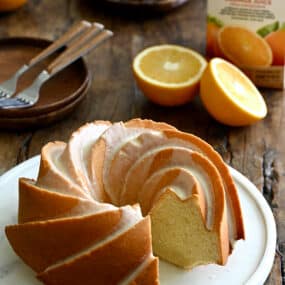 A Glazed Orange Pound Cake with one slice missing on a round serving plate next to an orange and a carton of Florida's Natural orange juice