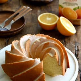 A Glazed Orange Pound Cake with one slice missing on a round serving plate next to an orange and a carton of Florida's Natural orange juice