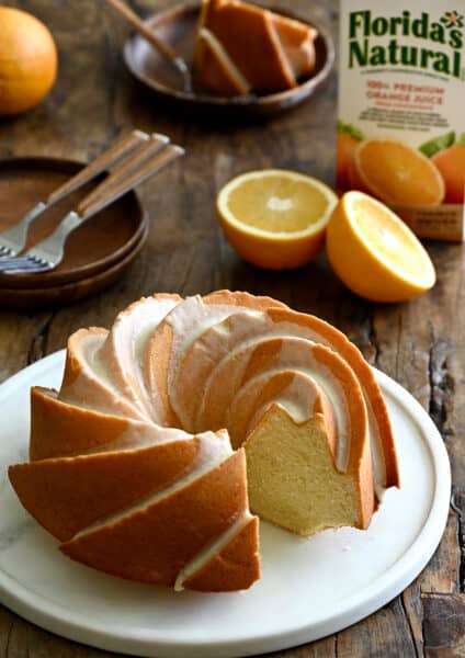 A Glazed Orange Pound Cake with one slice missing on a round serving plate next to an orange and a carton of Florida's Natural orange juice