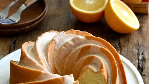 A Glazed Orange Pound Cake with one slice missing on a round serving plate next to an orange and a carton of Florida's Natural orange juice