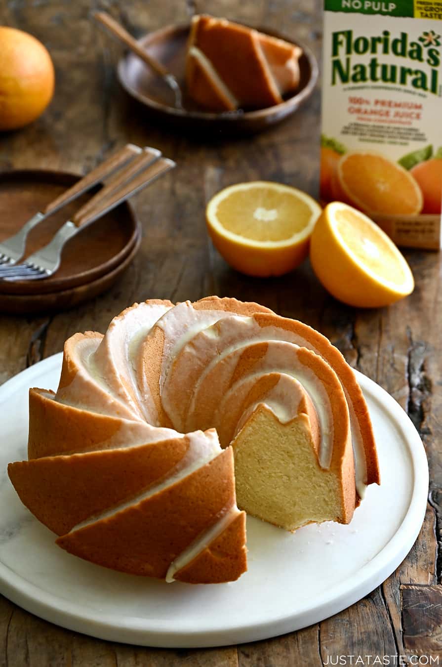 A Glazed Orange Pound Cake with one slice missing on a round serving plate next to an orange and a carton of Florida's Natural orange juice