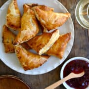 A top-down view of puff pastry brie and jam turnovers on a plate next to a plate containing green grapes, wine glasses filled with white wine, and a small bowl containing raspberry jam and a small spoon.