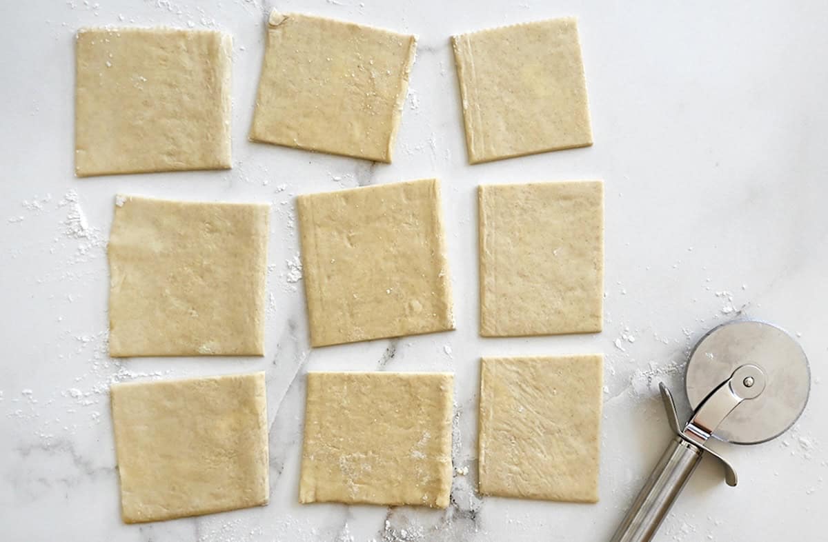 A sheet of store-bought puff pastry cut into nine squares next to a pizza cutter.