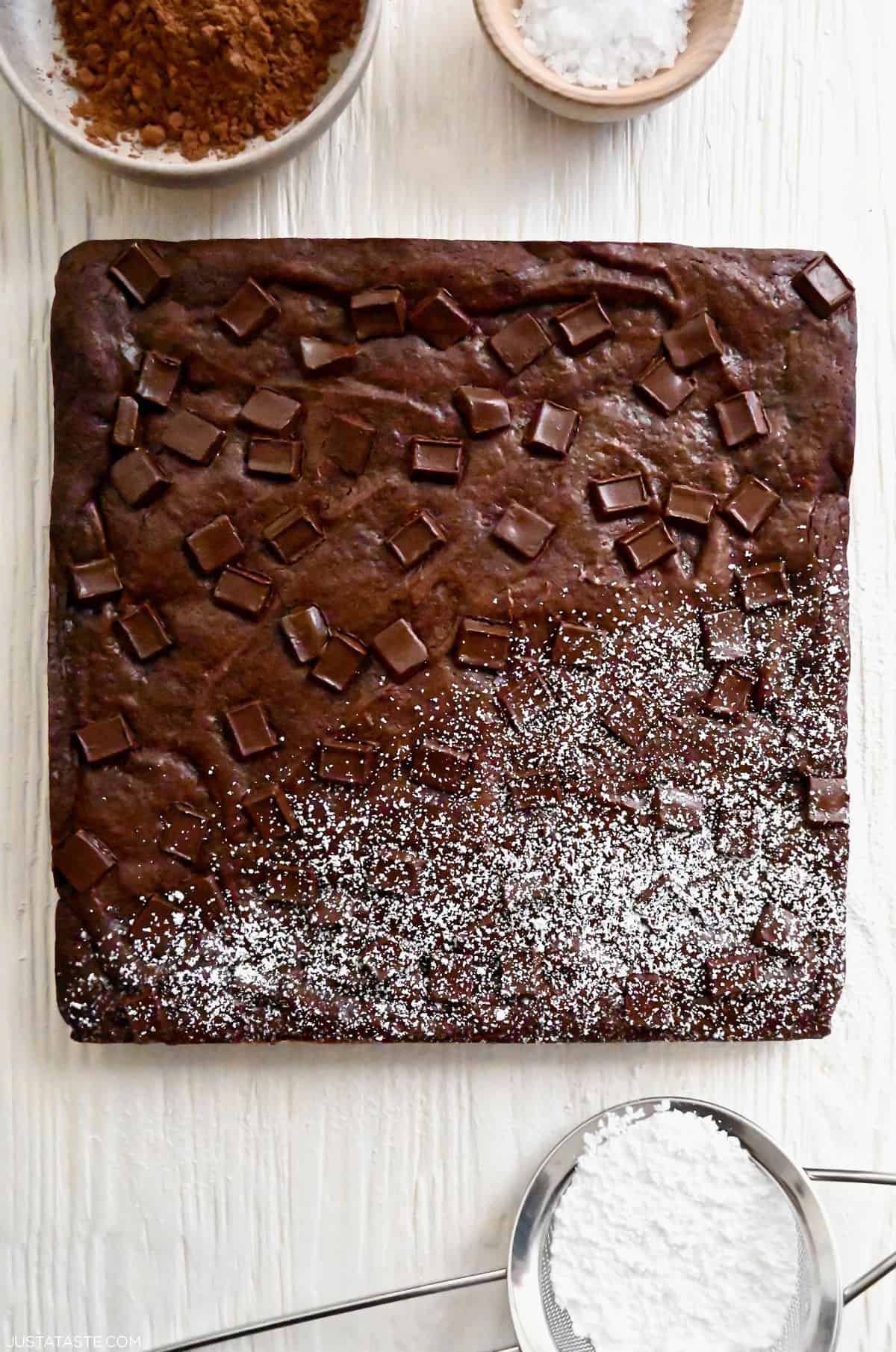 An uncut square brownie with chocolate chunks that's half dusted with powdered sugar.