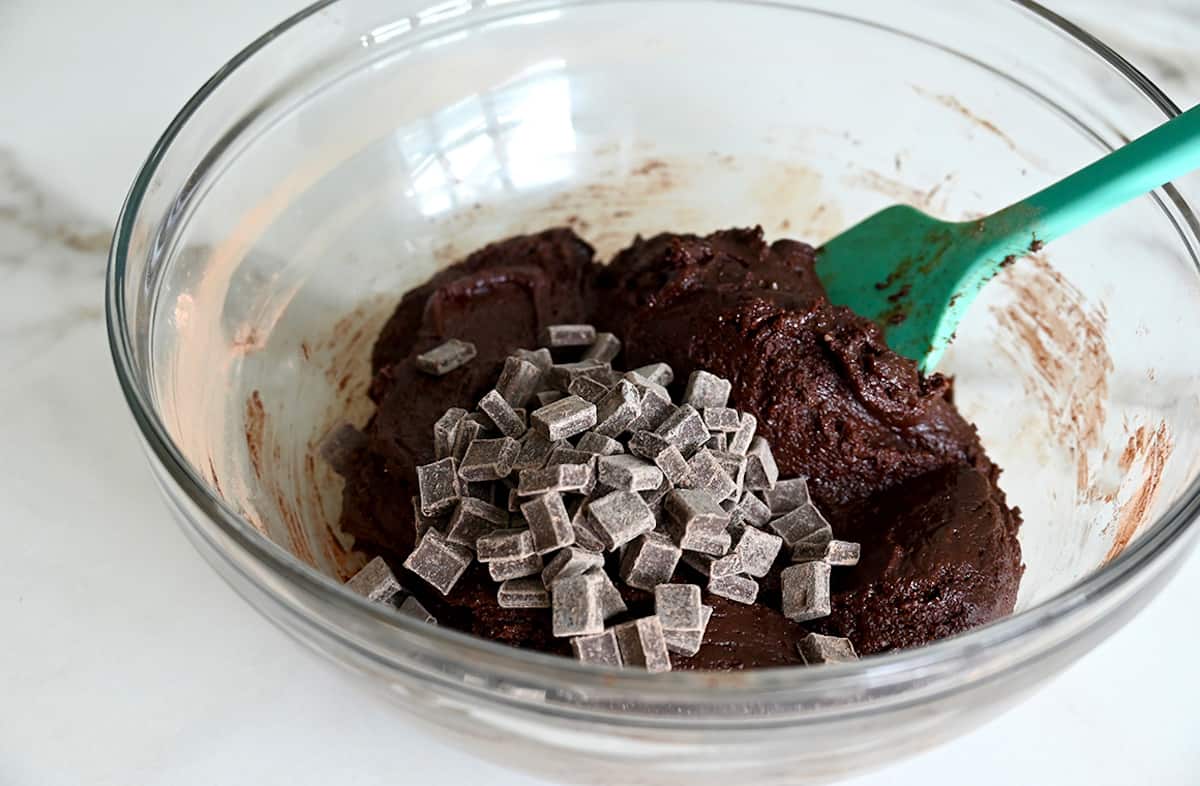 A teal spatula folds chocolate chunks into brownie batter in a glass bowl.