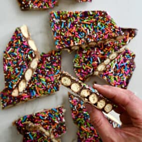 A hand holding a piece of pretzel bark topped with rainbow sprinkles