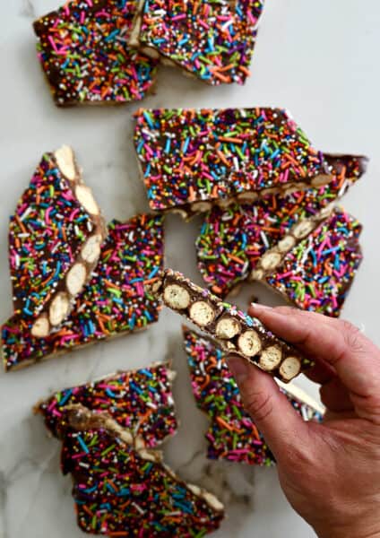 A hand holding a piece of pretzel bark topped with rainbow sprinkles