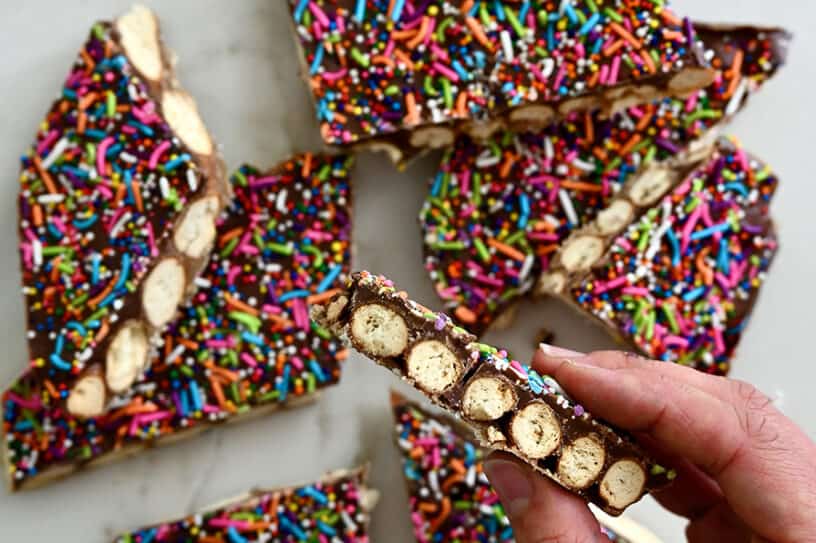 A hand holding a piece of pretzel bark topped with rainbow sprinkles