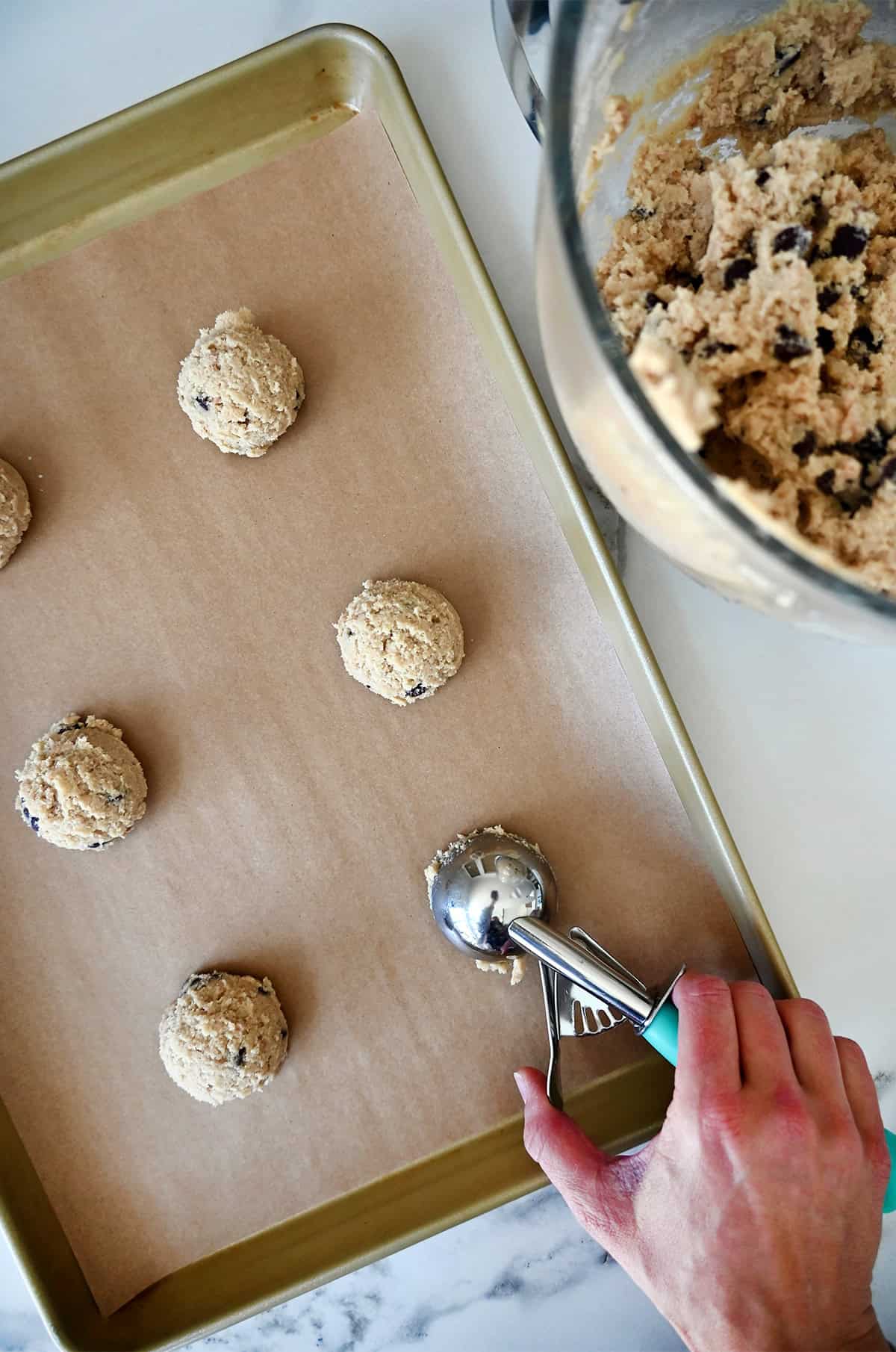 A hand holding a small cookie scoop adds mounds of cookie dough to a parchment paper-lined baking sheet. A bowl containing toffee cookie dough is nearby.