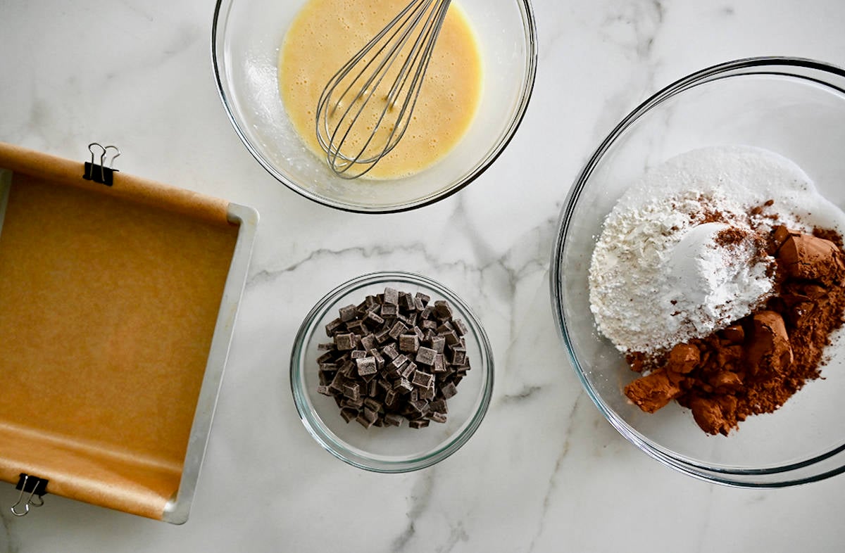 A square baking dish lined with parchment paper next to three glass bowls containing chocolate chunks, flour and cocoa powder, and a whisked egg.