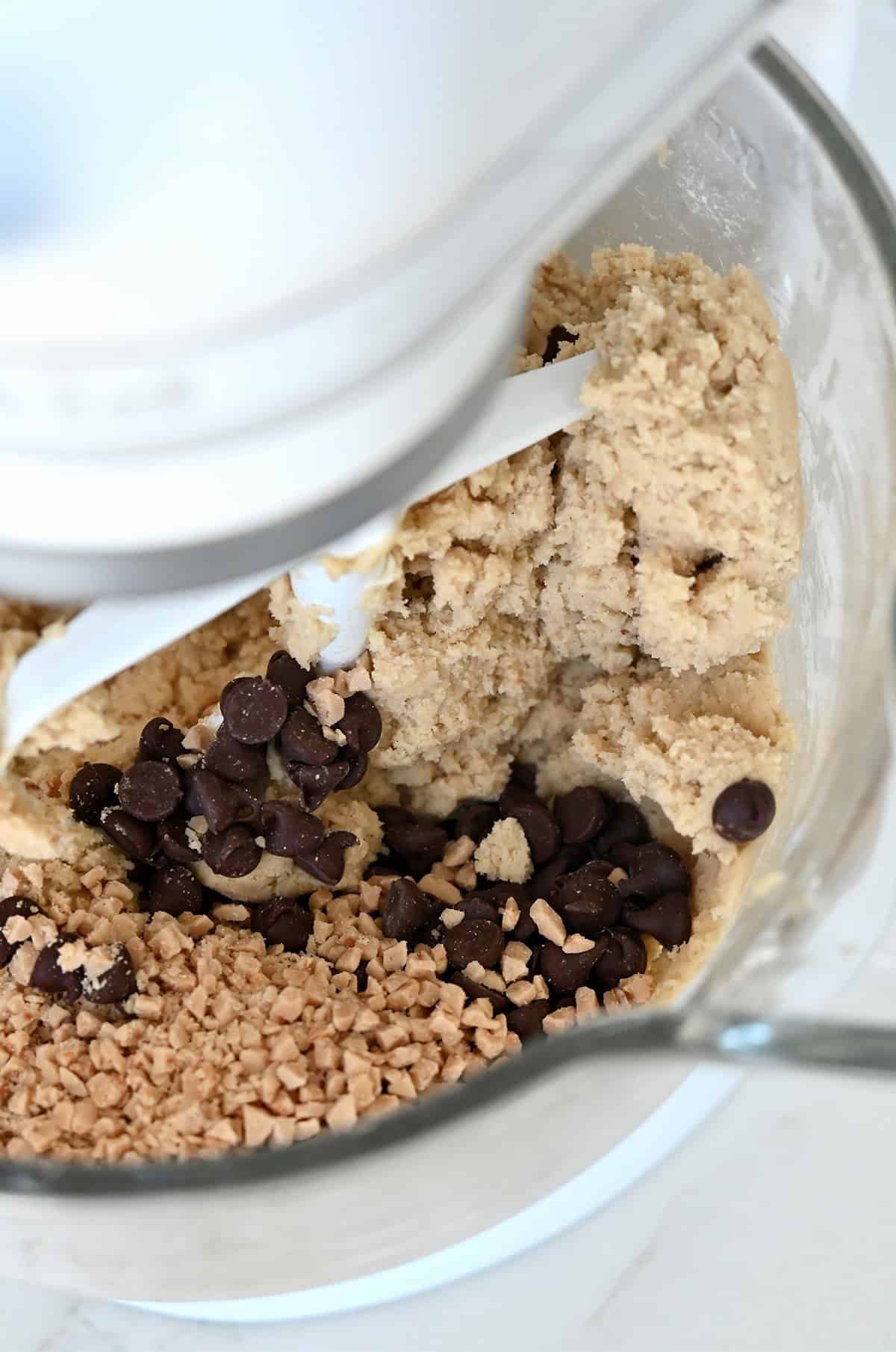 Toffee bits and chocolate chips atop cookie dough in the bowl of a stand mixer with the paddle attachment.