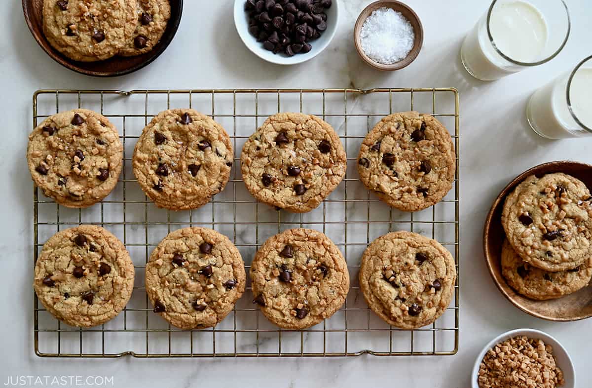 Two rows of toffee chocolate chip cookies cooling on a wire baking rack next to small bowls containing sea salt and chocolate chips, plus two glasses filled with milk.