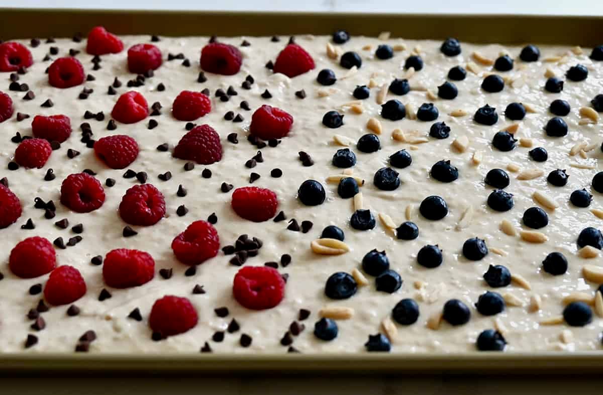 Berries, mini chocolate chips and nuts scattered atop pancake batter spread evenly on a baking sheet.