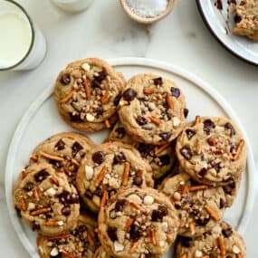 A top-down view of a white plate loaded with Kitchen Sink Cookies next to two glasses of milk and a small dish containing sea salt