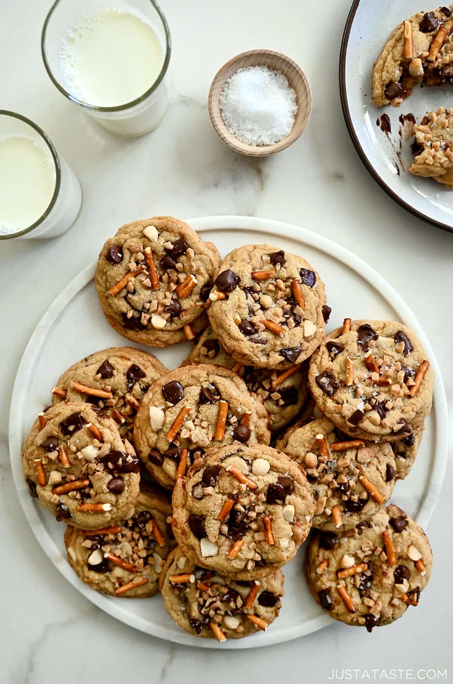 A top-down view of a white plate loaded with Kitchen Sink Cookies next to two glasses of milk and a small dish containing sea salt