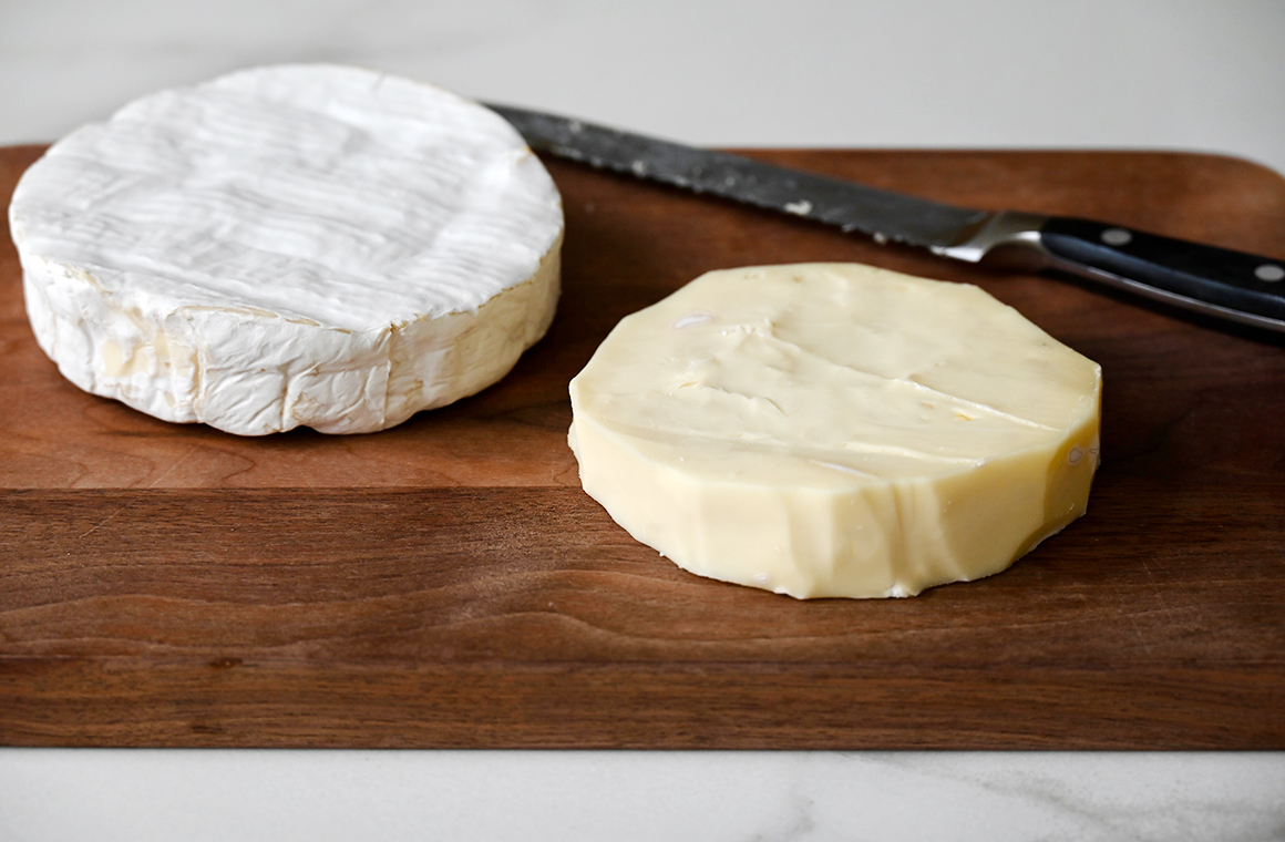 A wood cutting board containing two wheels of Brie cheese and a serrated knife