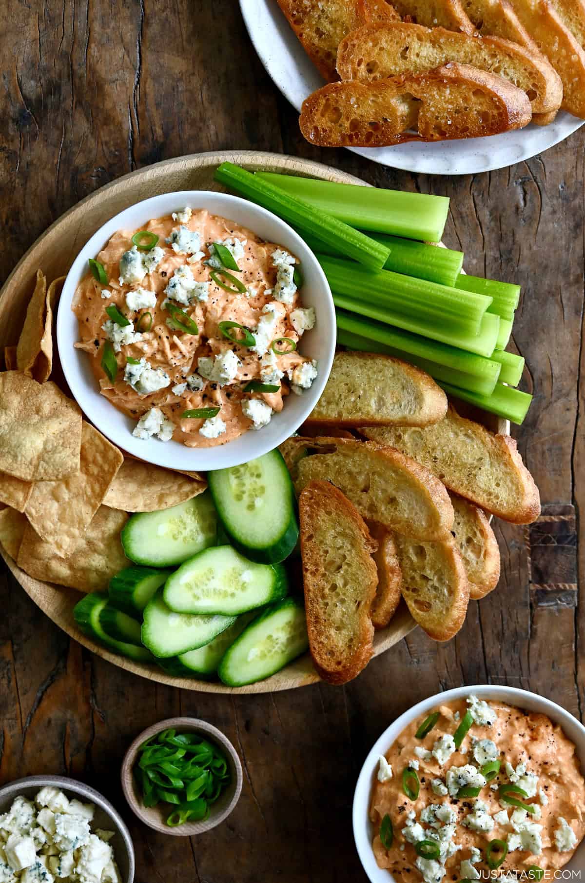 Buffalo chicken dip in a bowl garnished with blue cheese crumbles and sliced green onions on a platter surrounded by celery sticks, toasted baguette slices, cucumber slices and tortilla chips.