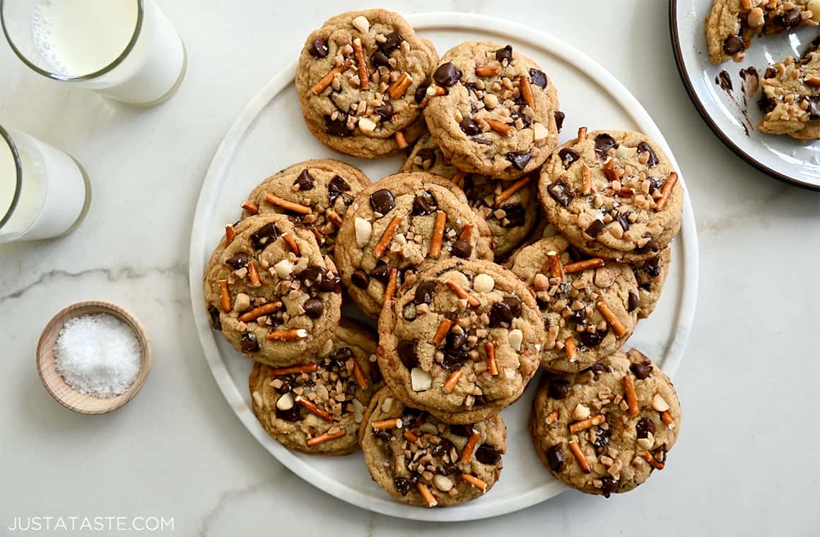 Kitchen Sink Cookies loaded with chocolate, chopped nuts, toffee bits and pretzels piled high on a plate next to two glasses containing milk