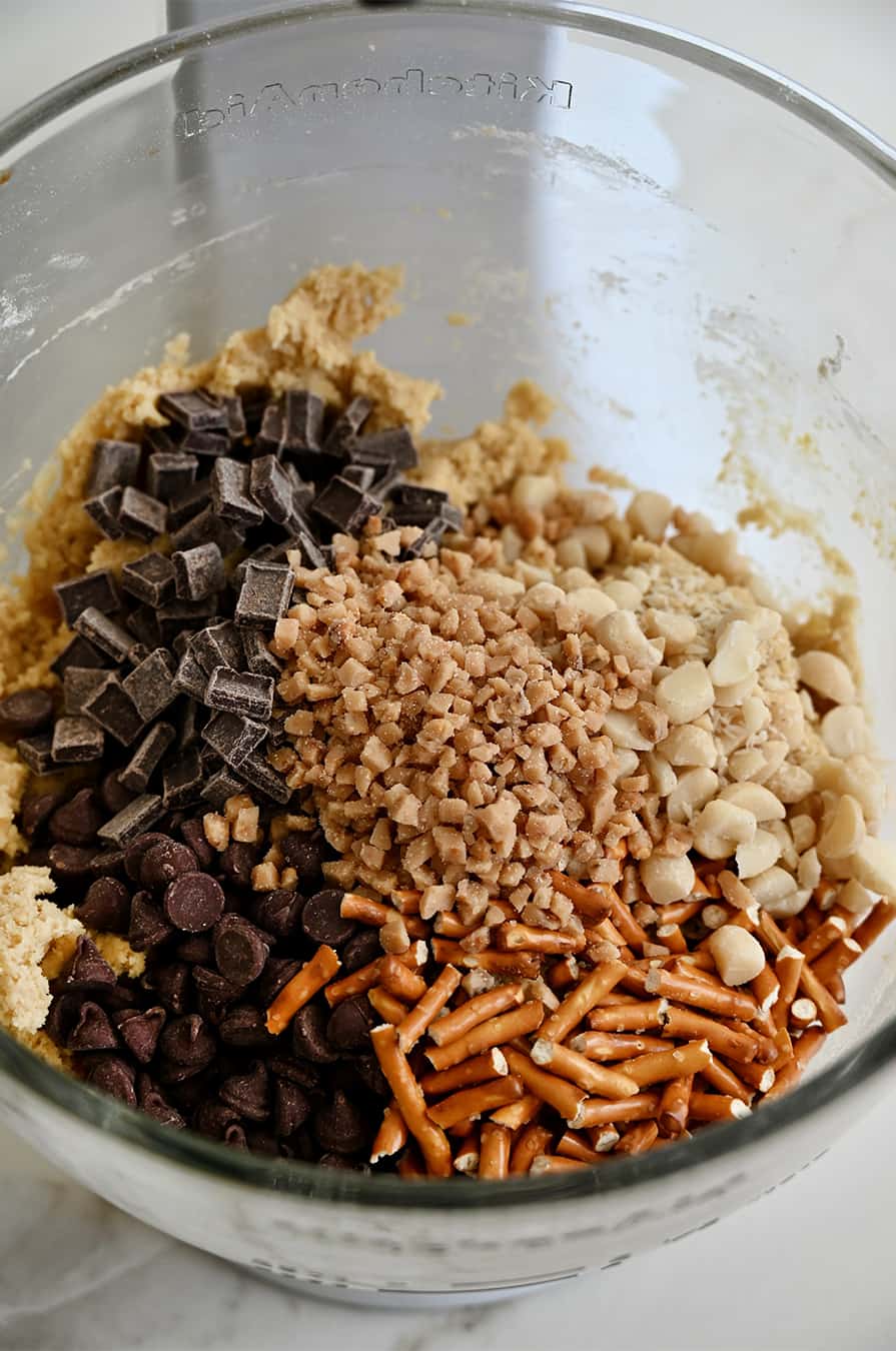 A clear bowl of a stand mixer containing toffee bits, crushed pretzel sticks, chocolate chunks, chopped macadamia nuts and semisweet chocolate chips