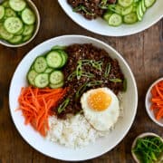 A top-down view of two bowls containing Korean beef, white rice, shredded carrots, sliced cucumbers and a fried egg.