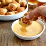 A hand holds a soft pretzel know just above a small bowl containing beer cheese dip with glasses of beer and more pretzel knots on a plate out of focus in the background.